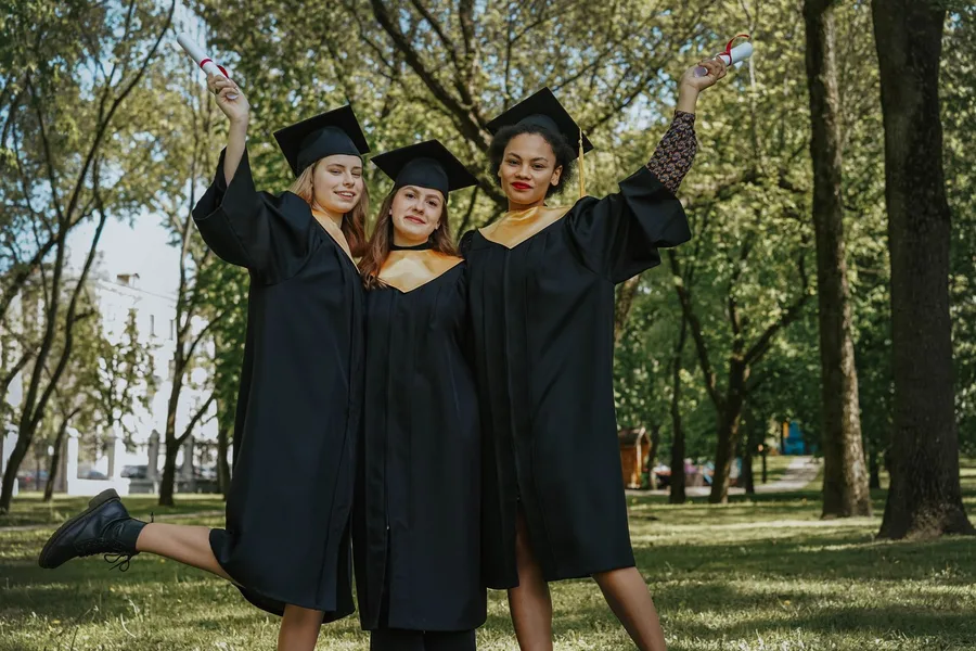 Three graduates in caps and gowns raising rolled diplomas outdoors