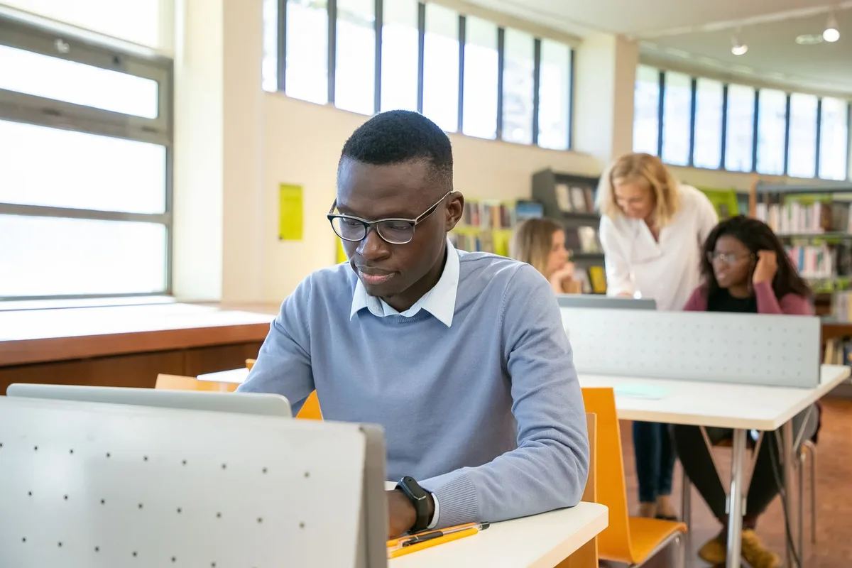 A college student working at a laptop in a university library while classmates study at nearby tables