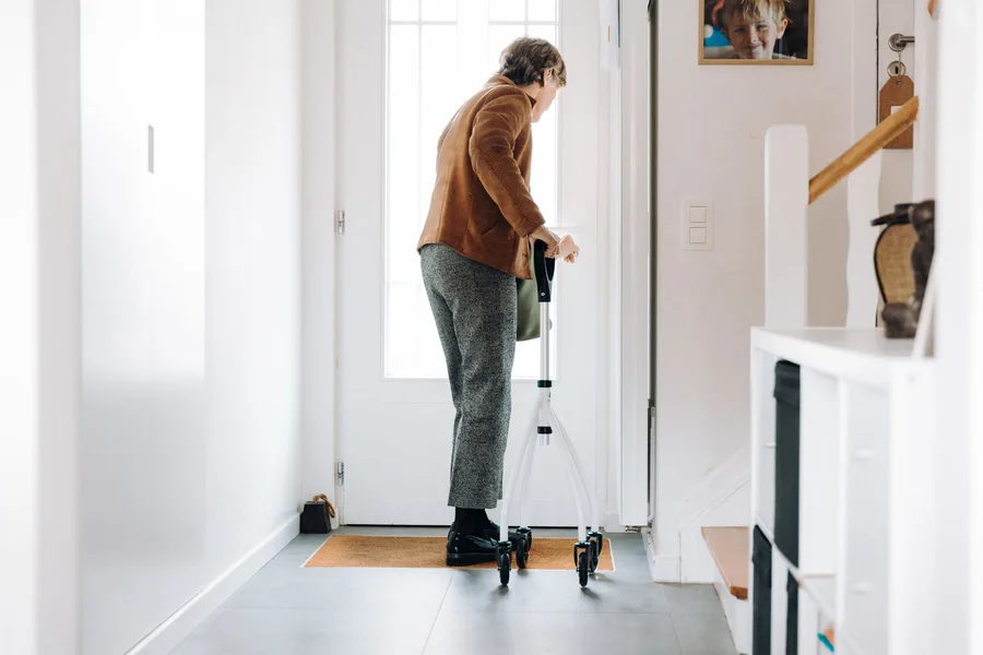 An older woman in a brown jacket stands at her own front door in a bright hallway, steadying herself on a wheeled walker