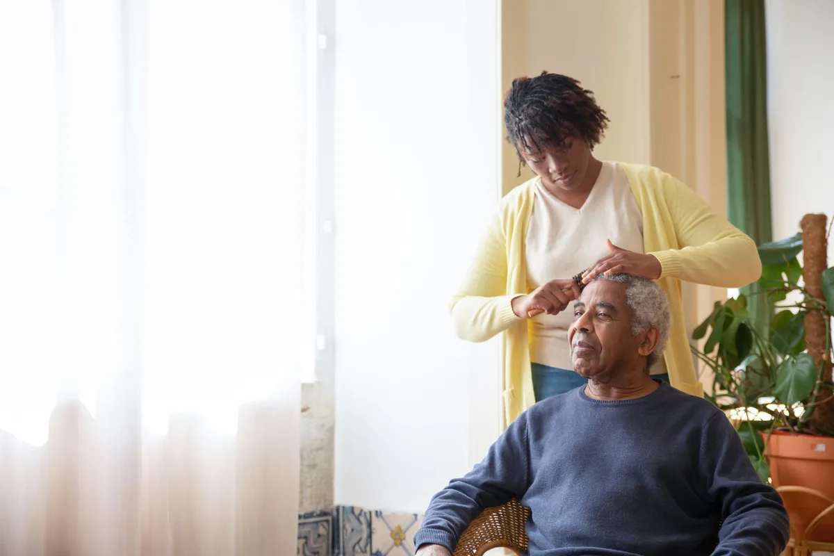 A caregiver stands behind an older man seated in a wicker chair by a bright window and combs his hair