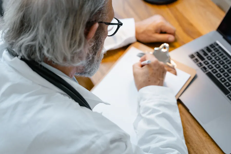 A doctor in a white coat with a stethoscope writes on a clipboard at a desk beside a laptop