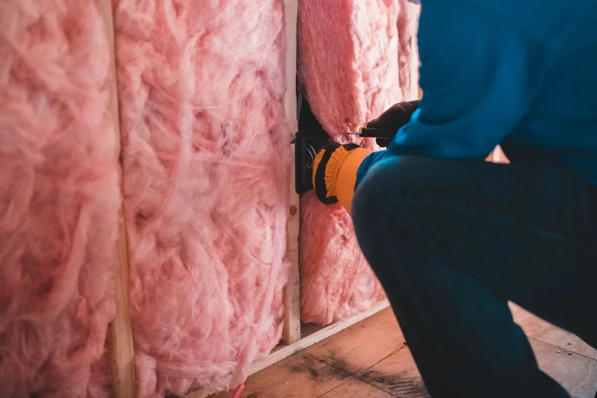 A worker in gloves fitting fiberglass insulation into a wall cavity