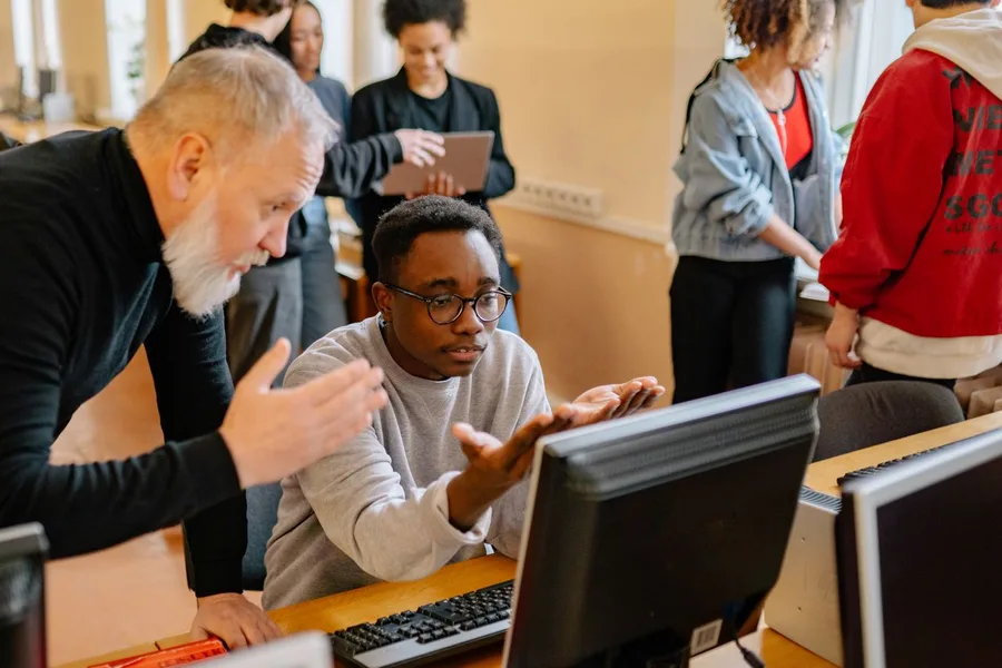 An older instructor helping a student at a monitor in a busy campus computer lab