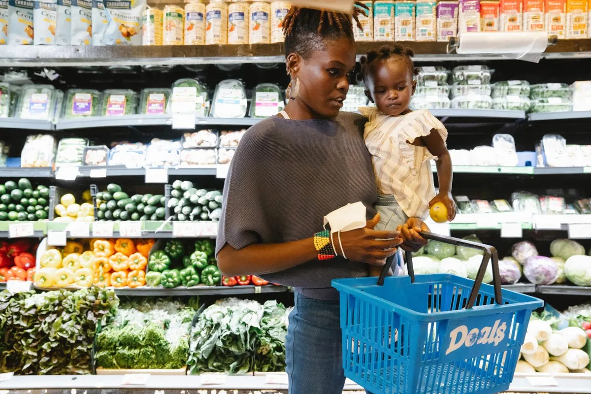 A parent holding a toddler while shopping in the fresh produce aisle of a grocery store, a shopping basket in hand