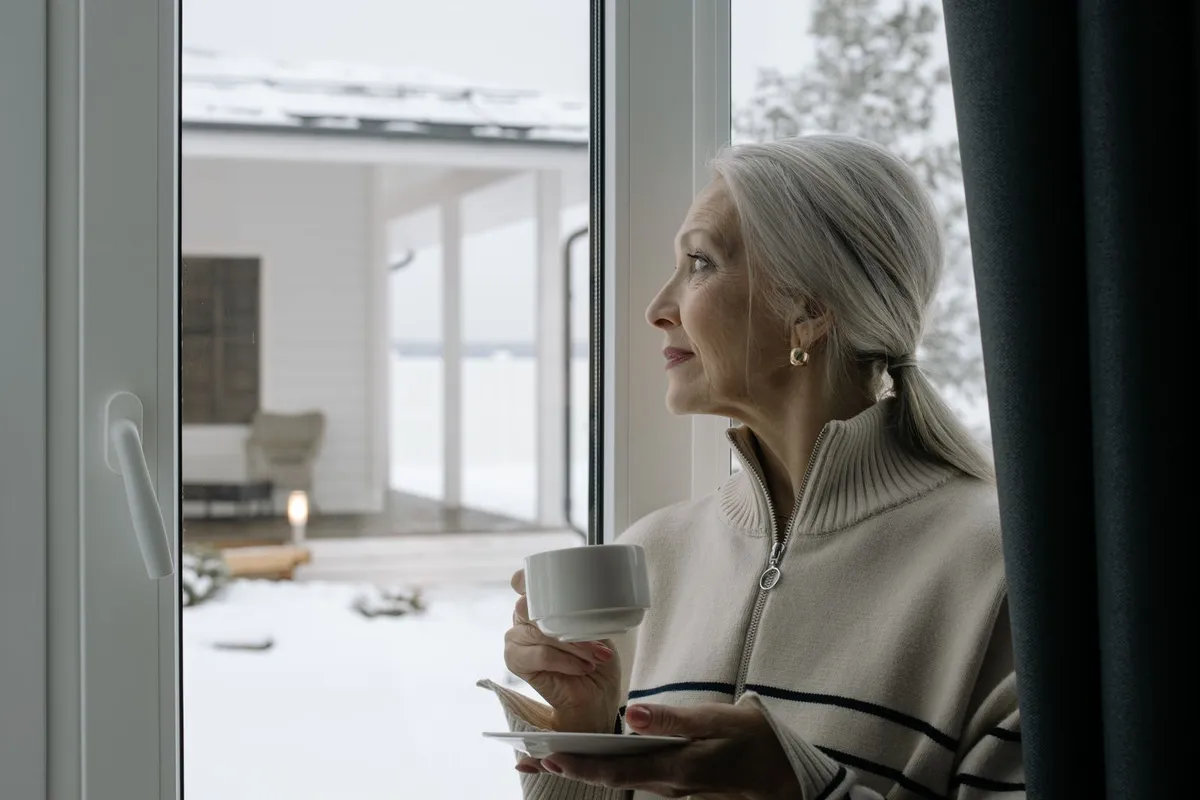 An older woman in a warm sweater holds a cup of tea and looks out a window at a snowy yard